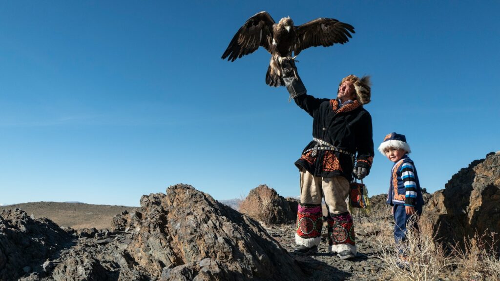 Kazakh eagle hunter holding Golden Eagle with young child standing beside him