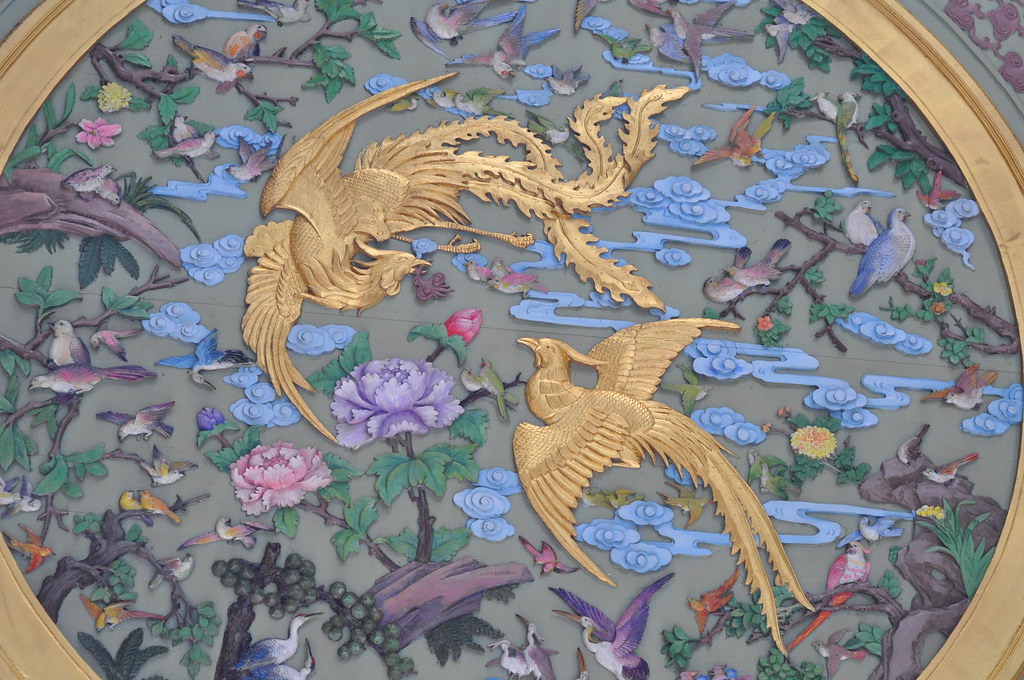 Golden Chinese phoenixes with spread wings fly amidst vibrant flowers and other birds in a detailed relief.