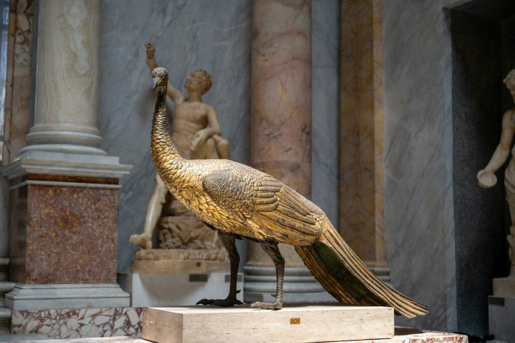 A detailed bronze peacock statue stands on a pedestal in a museum setting.