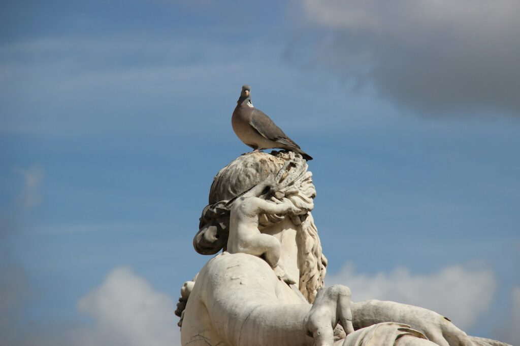 A pigeon stands atop a weathered stone statue, against a partly cloudy sky.