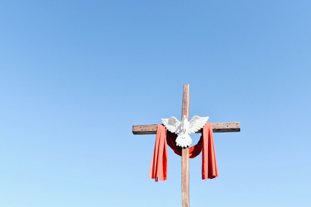 A wooden cross is adorned with a white dove and draped with red cloth against a clear sky.
